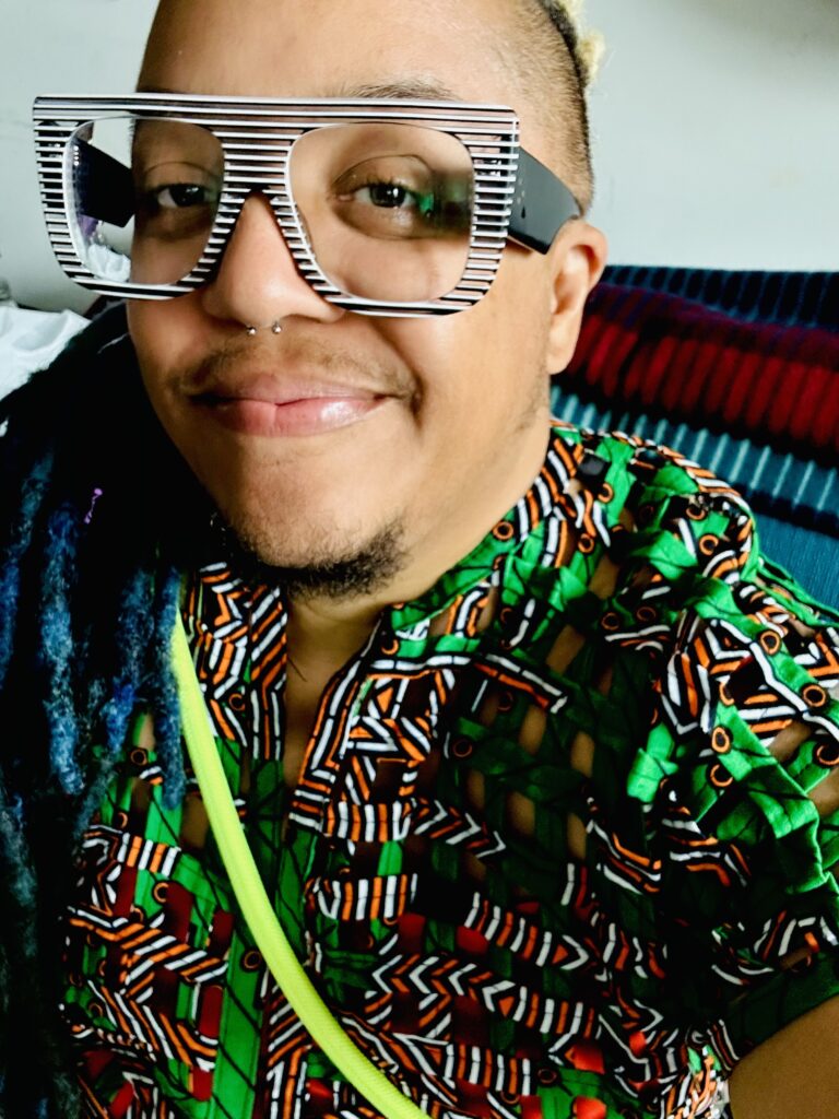 Lawyers Against Transphobia National Conference 2025: Educate! Advocate! Agitate! - Meet our facilitators. Pictured - Syrus Marcus Ware. Syrus, a Black man with blue locs, is pictured in chunky glasses and a colourful top, smiling.
